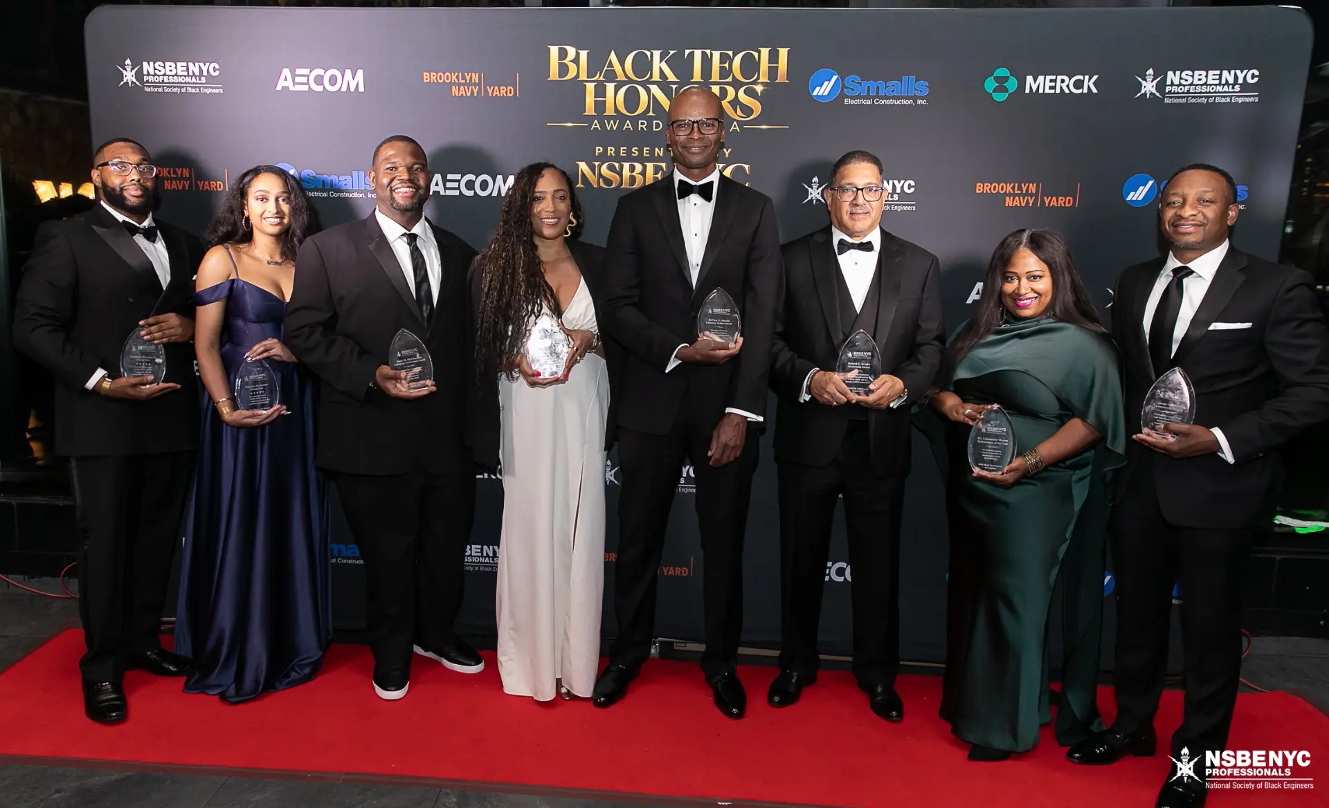 The eight 2026 Black Tech Honors award recipients in formal attire holding their crystal awards on the red carpet in front of the Black Tech Honors sponsor backdrop
